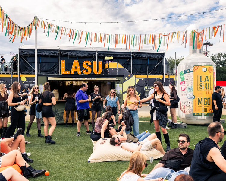 LA SU Festival-Bar mit Leuchtschrift und übergroßer MGO-Flasche, Gäste feiern auf der Wiese