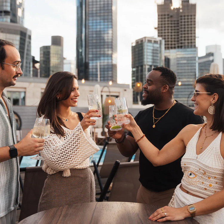 Vier Freunde stoßen mit LA SU Gläsern auf Dachterrasse vor Frankfurter Skyline an
