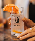Hand hält LA SU Glas mit Drink und Orangenscheibe