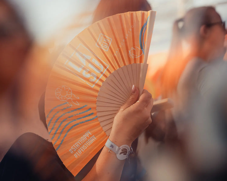 Person hält LA SU Fächer mit #StayHappy-Aufdruck bei Sonnenuntergang